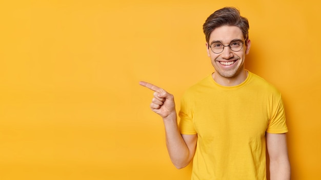 La toma horizontal de un hombre adulto feliz señala con el dedo índice hacia afuera y muestra un anuncio contra el fondo amarillo del estudio lleva anteojos redondos, una camiseta informal demuestra una promoción Mire esto, por favor