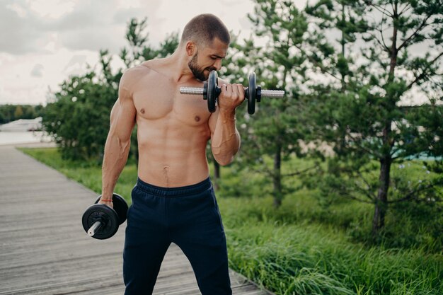 La toma horizontal de un hombre musculoso motivado hace ejercicio con una barra demuestra que el poder de la masculinidad tiene un cuerpo fuerte y se para sin camisa al aire libre se esfuerza por levantar peso con esfuerzo hace curl de bíceps