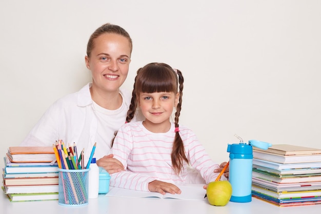 Toma horizontal de una mujer sonriente sentada con su hija en el escritorio aislada sobre fondo blanco haciendo la tarea juntos posando rodeada de libros mirando la cámara con sonrisas