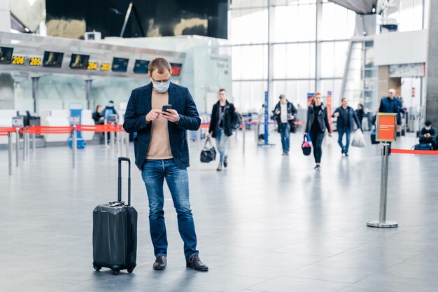 La toma horizontal de poses de hombre en un aeropuerto abarrotado usa teléfonos celulares para verificar el tiempo de vuelo en línea se encuentra cerca de la maleta usa una máscara médica durante la crisis del coronavirus Concepto de viaje de atención médica