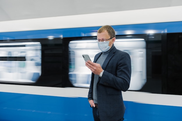 La toma horizontal de un viajero masculino usa el transporte público para viajar, usa una máscara médica para protegerse del coronavirus o covid-19, espera el tren, usa el teléfono móvil, envía mensajes de texto en el chat