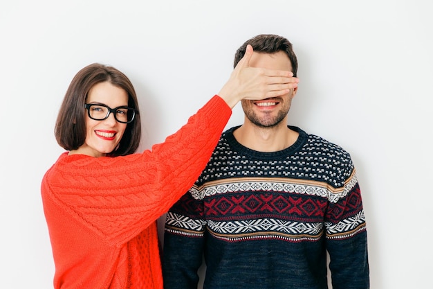 Toma interior de una atractiva morena con expresión positiva que cubre los ojos de los hombres, usa suéteres de invierno, se paran juntos contra un fondo blanco, diviértete. Eso es una sorpresa para ti, no mires.