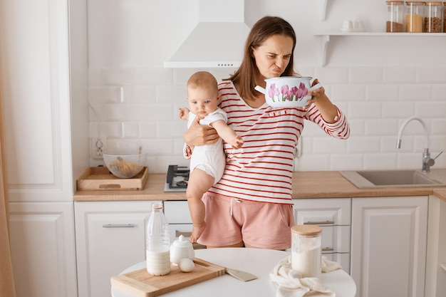 Toma interior de una atractiva mujer de cabello oscuro con camisa a rayas y una pose corta en la cocina con su hija pequeña sosteniendo una olla y oliendo el olor de la cama