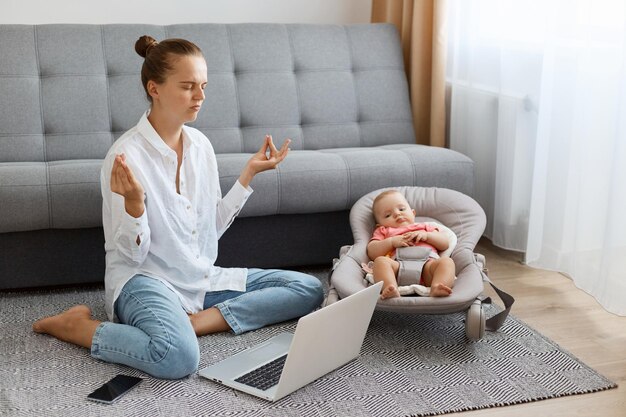 Toma interior de una atractiva mujer cansada con peinado de moño con camisa blanca y jeans sentada en el suelo frente a la computadora portátil y cuidando al bebé haciendo yoga para calmarse