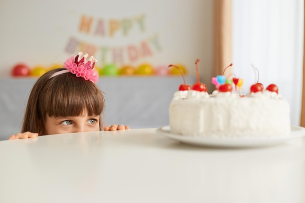 Toma interior de una curiosa y encantadora niñita escondida detrás de la mesa, espiando, mirando, quiere ver un delicioso pastel de cumpleaños, decoraciones festivas en el fondo.