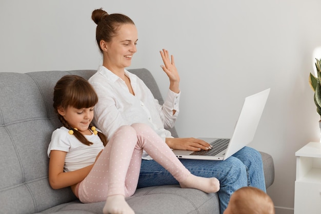 Toma interior de familia feliz, madre e hija sentadas en un cómodo sofá y conversando en línea a través de una computadora portátil, mujer joven agitando la mano a la cámara de la computadora portátil.