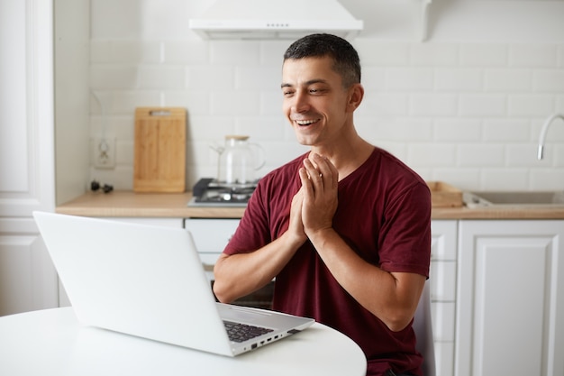 Toma interior de un hombre independiente guapo extremadamente feliz, trabajando en línea en casa en la cocina, teniendo buenas noticias del empleador, aplaudiendo, regocijándose de su éxito, vistiendo ropa informal.