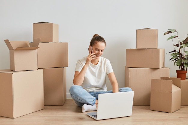 Toma interior de una joven adulta positiva con una camiseta blanca sentada en el suelo cerca de cajas de cartón con cosas, trabajando en una computadora portátil y hablando por teléfono con un socio comercial.