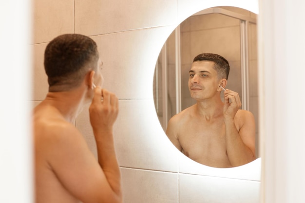 Toma interior de un joven adulto moreno después de tomar una ducha, reflejo en el espejo del hombre atractivo desnudo haciendo procedimientos de higiene en el baño, limpiando sus oídos con un hisopo de algodón.