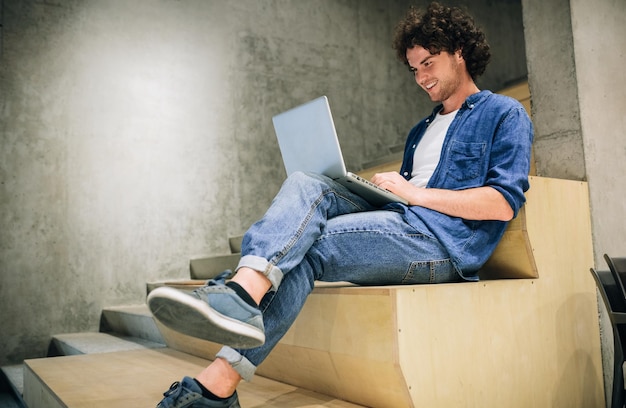 Toma interior de un joven con cabello rizado usando una computadora portátil para chatear en línea con amigos navegando por Internet sentado en las escaleras en el muro de concreto Hombre sincero leyendo y escribiendo en su computadora portátil