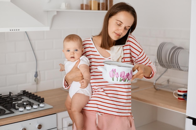 Toma interior de una joven madre ocupada parada con una hija pequeña en las manos y hablando por teléfono sosteniendo una olla con sopa y mirando adentro usando una camisa casual a rayas