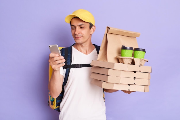 Toma interior de un joven mensajero con gorra y camiseta blanca sosteniendo pizza y café en vasos desechables usando un teléfono celular para comunicarse con un cliente aislado en un fondo morado