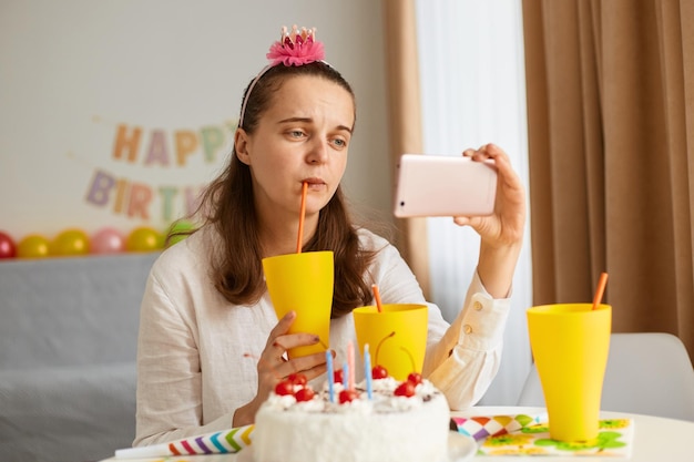 Toma interior de una joven triste con camisa blanca celebrando un cumpleaños sola bebiendo una bebida de una taza amarilla sosteniendo un teléfono inteligente y haciendo una videollamada o una transmisión en vivo