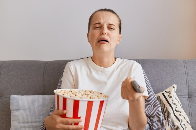 Toma interior de una joven triste y disgustada con una camiseta blanca sentada en un sofá con palomitas de maíz y control remoto viendo una película romántica o ve un momento trágico en la serie