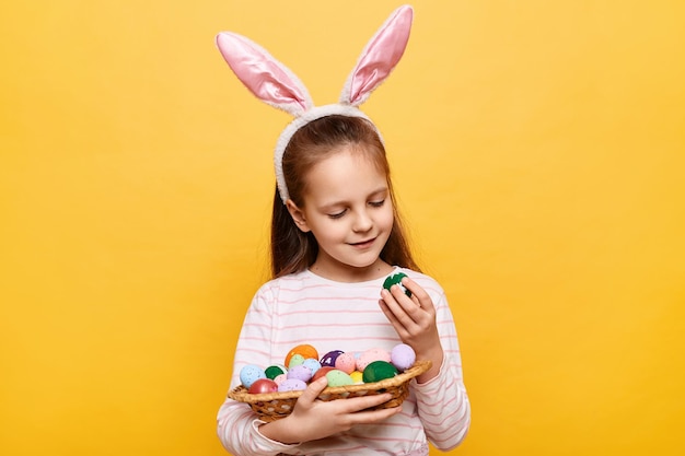 Toma interior de una linda niña positiva con orejas de conejo con una canasta de huevos de colores mirando patrones en símbolos de primavera posando aislados en un fondo amarillo