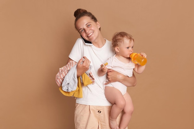 Toma interior de una madre sonriente y optimista con una hija pequeña posando aislada sobre una mujer de fondo marrón vestida con una camiseta blanca hablando por teléfono móvil expresando felicidad