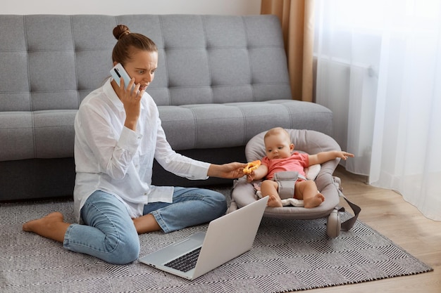 Toma interior de una mujer atractiva con una camiseta blanca sentada cerca de la tos con una hija pequeña en una mecedora y trabajando en una laptop hablando por teléfono con un socio comercial