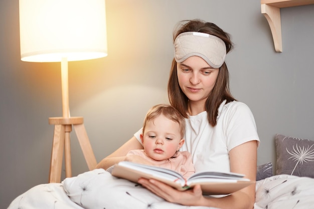 Toma interior de una mujer de cabello oscuro posando con su hija en el dormitorio madre leyendo un libro a su bebé mientras se sientan juntos en la cama antes de irse a dormir