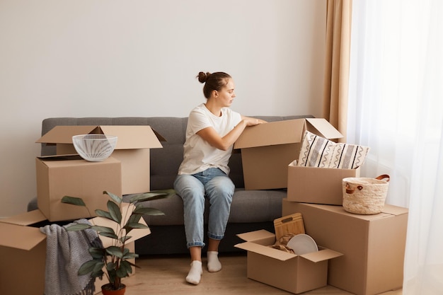 Toma interior de una mujer caucásica concentrada con camiseta blanca y jeans sentada en un sofá en una nueva casa o apartamento después de mudarse, desempacando sus pertenencias de cajas de cartón.