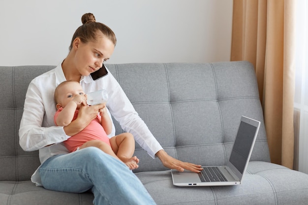 Toma interior de una mujer caucásica molesta con un peinado de moño con camisa blanca y jeans sosteniendo a su bebé y hablando por teléfono mientras trabaja en una laptop sentada en un sofá