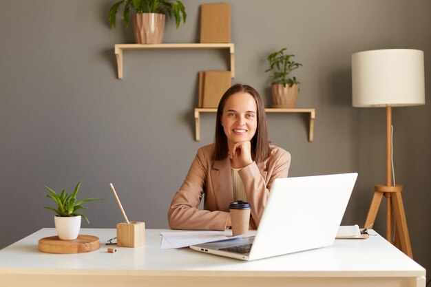 Toma interior de una mujer caucásica sonriente y satisfecha independiente o gerente con chaqueta beige sentada en la mesa cerca de la computadora portátil mirando la cámara sosteniendo la barbilla