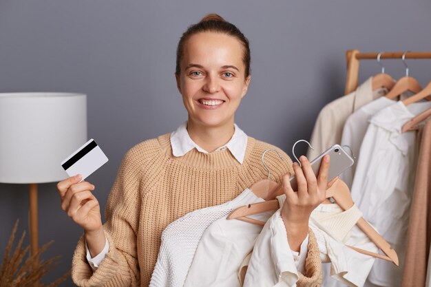 Toma interior de una mujer hermosa y atractiva sonriente adicta a las compras sosteniendo ropa en perchas en las manos de pie con teléfono celular y tarjeta de crédito pagando en línea por nuevos atuendos