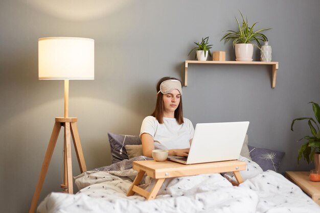 Toma interior de una mujer independiente seria y concentrada que usa máscara para dormir sentada con una computadora portátil en la cama en casa trabajando en línea después de despertarse por la mañana