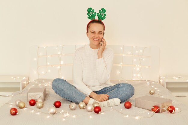 Toma interior de una mujer optimista sonriente con un suéter de estilo casual y jeans, posando en un dormitorio blanco, sentada en la cama y hablando por teléfono celular, felicitando a los familiares por el año nuevo.