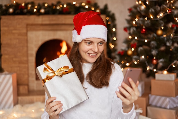 Toma interior de mujer positiva feliz vestida con suéter blanco y sombrero de santa claus, posando junto a la chimenea y el árbol de Navidad, teniendo una videollamada, mostrando su regalo de año nuevo a la cámara del gadget.
