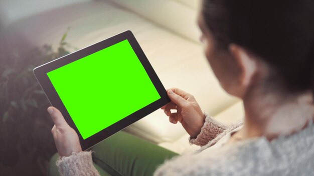 Toma interior de una mujer que usa tablet pc con pantalla verde sentada en un sofá blanco con orientación horizontal