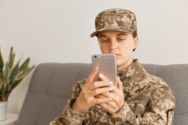 Toma interior de una mujer soldado concentrada con uniforme de camuflaje y gorra sosteniendo un teléfono celular y navegando por Internet o revisando el correo electrónico militar trabajando