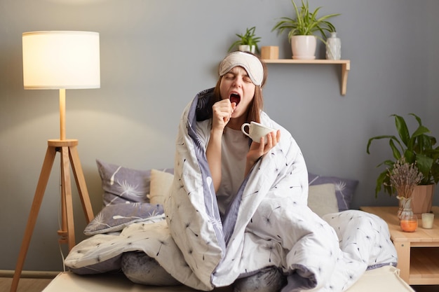 Toma interior de una mujer soñolienta envuelta en una manta después de despertarse sosteniendo una taza de café cubriendo la boca con la mano se siente insomne bebiendo una bebida energética