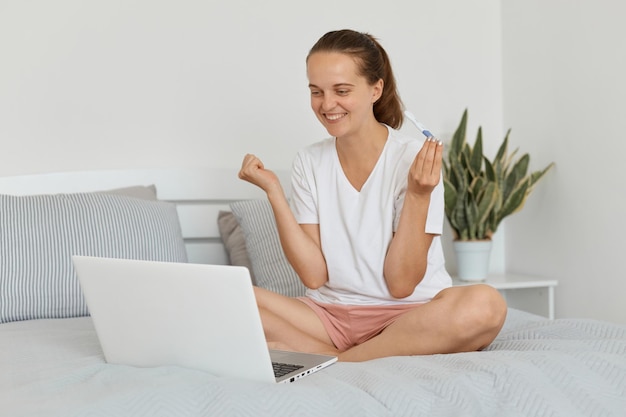 Toma interior de una mujer sonriente sentada en la cama y hablando a través de una videollamada en una computadora portátil contando sobre su embarazo alardeando del resultado mirando el dispositivo mostrando los puños apretados y celebrando buenas noticias