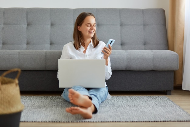 Toma interior de una mujer sonriente vestida con camisa blanca y jeans trabajando en línea a través de una computadora portátil en casa, tomando un descanso, revisando las redes sociales, correo electrónico, usando un teléfono inteligente, sentada en el piso en una habitación luminosa.