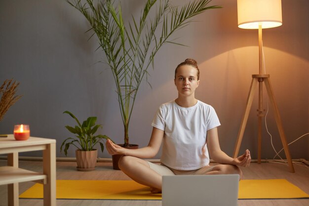 Toma interior de una mujer tranquila y atractiva con camiseta blanca practicando yoga y estirando el cuerpo usando una computadora portátil para la clase en línea sentada en una pose de loto con los ojos cerrados meditando relajante