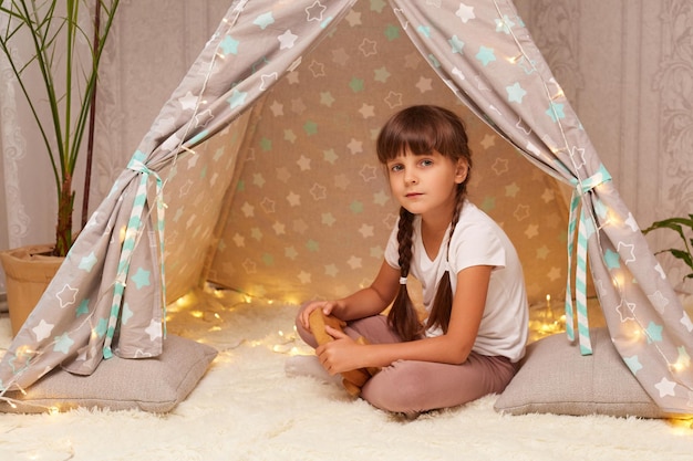 Toma interior de una niña seria con trenzas con camiseta blanca posando en wigwam mirando a la cámara con expresión tranquila triste de jugar sola en casa