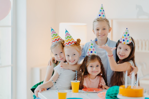 Toma interior de niños felices que celebran la fiesta con confeti cayendo, usan sombreros de fiesta de cono, posan junto a la mesa festiva con pasteles
