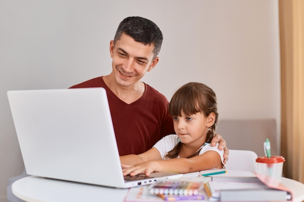 Toma interior del padre con una camiseta granate haciendo la tarea con la hija teniendo una lección en línea en la computadora portátil, el padre y la niña aprenden en casa, tienen una clase web en la computadora
