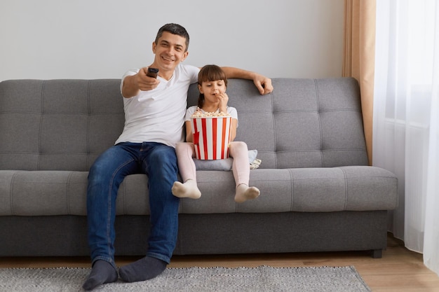 Toma interior de padre e hija pequeña viendo la televisión o una película sentada en el sofá con una canasta de palomitas de maíz papá sosteniendo el control remoto expresando emociones positivas