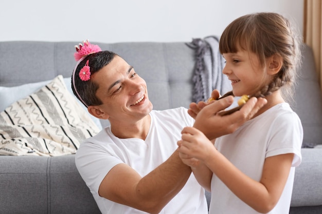 Toma interior de un padre feliz tocando el cabello de sus hijas mientras se sienta en el suelo cerca del sofá en casa, un hombre moreno mirando a su hijo con amor y una sonrisa encantadora