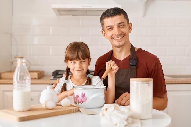 Toma interior de padre sonriente y linda hija posando en la cocina mientras está sentado a la mesa con productos para hacer pasteles caseros, familia mirando a cámara con expresión facial positiva.