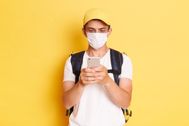 Toma interior del repartidor con camiseta de gorra amarilla y máscara protectora con mochila térmica para alimentos usando un teléfono celular mientras está de pie aislado en fondo amarillo