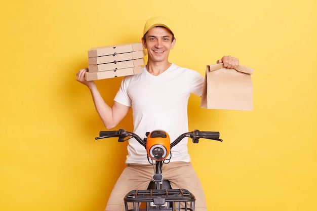 Toma interior de un repartidor montando en bicicleta y sosteniendo paquetes de papel con comida para llevar con camiseta y gorra mirando a la cámara con una sonrisa aislada sobre fondo amarillo