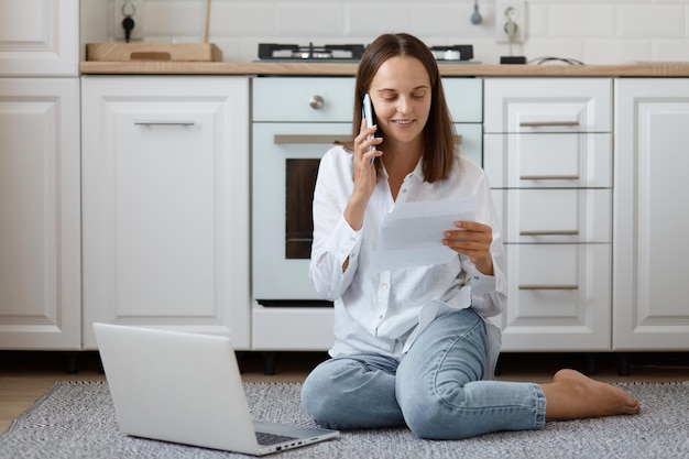 Toma interior de sonriente mujer positiva vestida con camisa blanca y jeans, sosteniendo una hoja de papel y hablando por teléfono, paga facturas de servicios públicos, trabaja en la computadora, posando en el piso de la cocina.