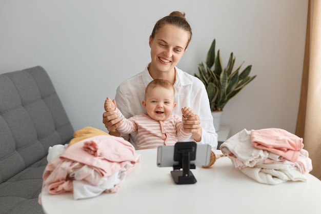 Toma en interiores de una bloguera sonriente que transmite una transmisión en vivo mientras está sentada a la mesa con su bebé y juega con ella, usando el teléfono móvil para grabar videos o transmitir en vivo, el blog de mamá.