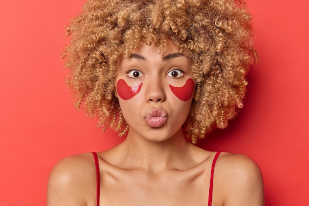 La toma en interiores de una encantadora mujer de pelo rizado sopla aire beso mantiene los labios redondeados tiene un estado de ánimo romántico aplica parches de hidrogel debajo de los ojos para reducir la hinchazón aislado sobre fondo rojo. Concepto de belleza