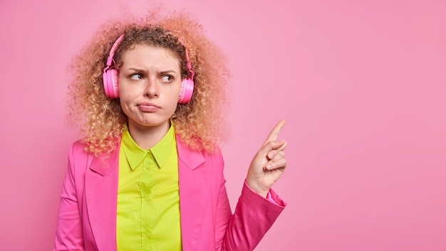 La toma en interiores de una joven de pelo rizado disgustada indica que en la esquina superior derecha demuestra algo desagradable vestido formalmente escucha música a través de auriculares aislados sobre una pared rosa