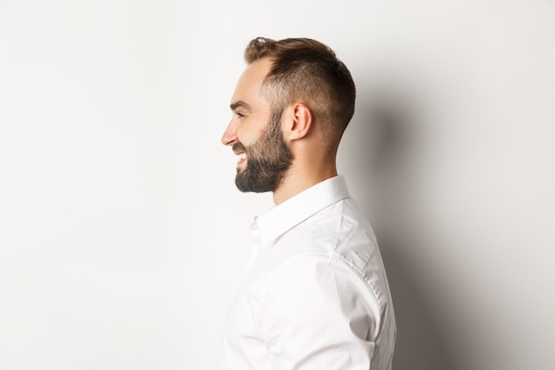 Toma de perfil de primer plano de hombre guapo con barba mirando a la izquierda y sonriendo, de pie contra el fondo blanco.