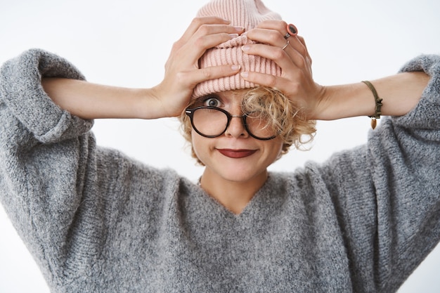 Toma de primer plano de la hermosa moderna y elegante mujer hipster de moda tirando gorro en la frente sonriendo alegremente y mirando al frente con mirada despreocupada feliz tomados de la mano en la cabeza sobre la pared blanca