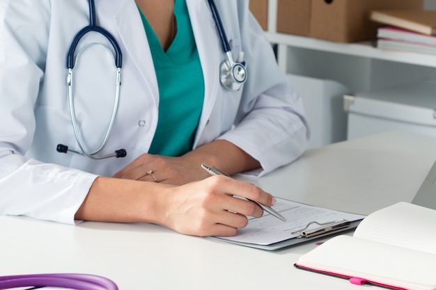 Toma de primer plano de las manos de la doctora en medicina llenando el formulario de registro del paciente. Lugar de trabajo del médico. Concepto médico y sanitario.